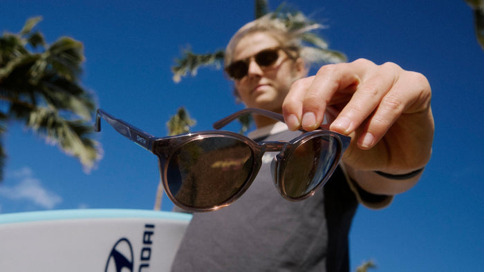 Molly Picklum tenant une planche de surf sous un bras et tenant une paire de lunettes de soleil dans l’autre main. 