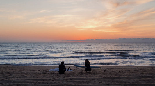The silhouettes of two people sitting on a beach at sunset.