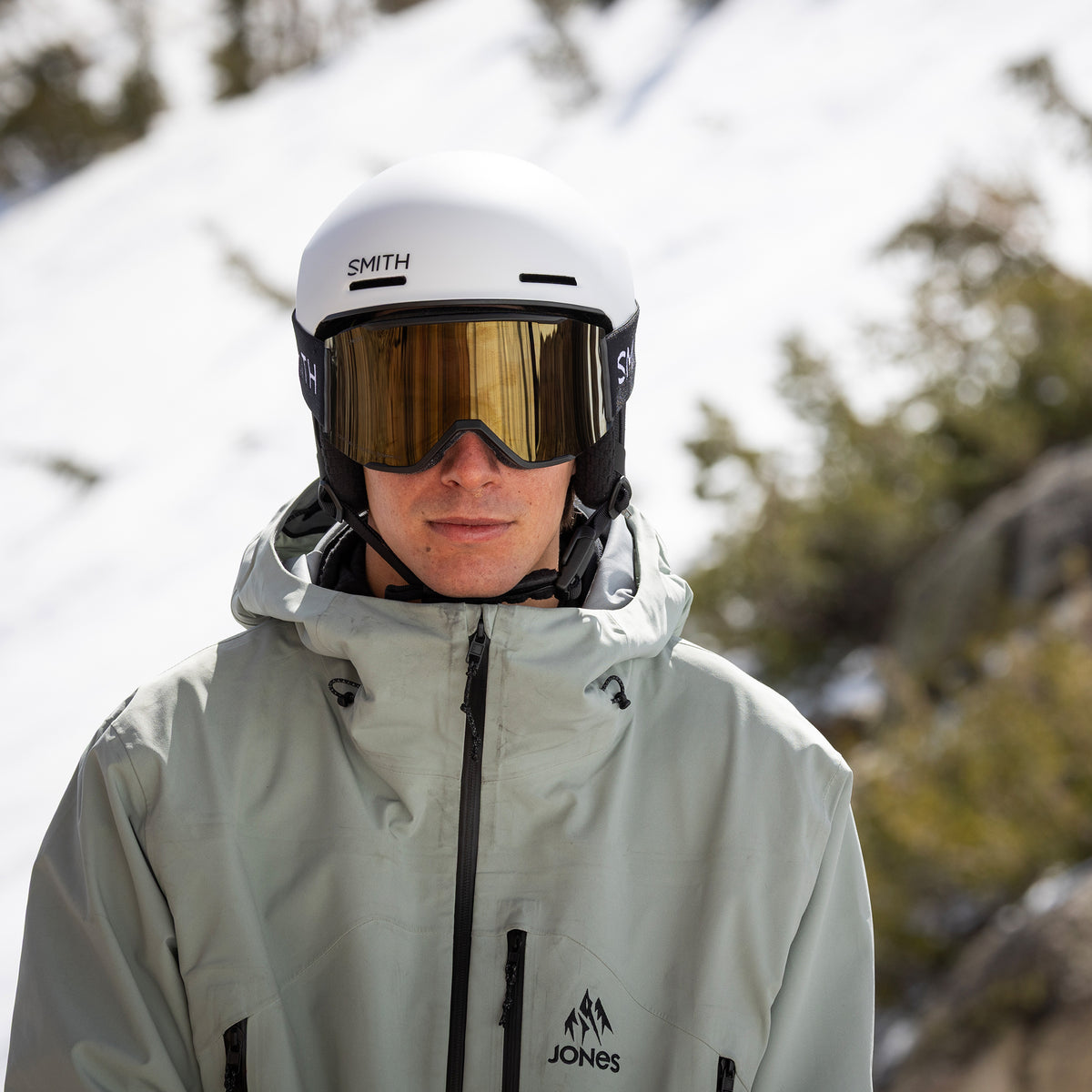 A person in a snowy outdoor setting wearing a white Smith helmet, gold-tinted ski goggles, and a light gray snowboard jacket with a black zipper and logo.